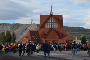 Große rote Holzkirche, dahinter Berg, im Vordergrund Menschen